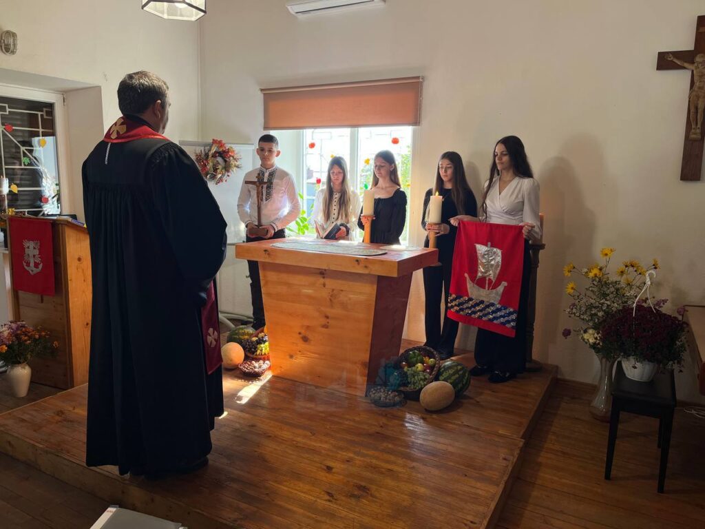 5 Jugendliche stehen hinter dem Altar, der mit Kürbissen und Wassermelonen geschmückt ist. Der Pfarrer steht links im Bild mit dem Rücken zum Fotografen.